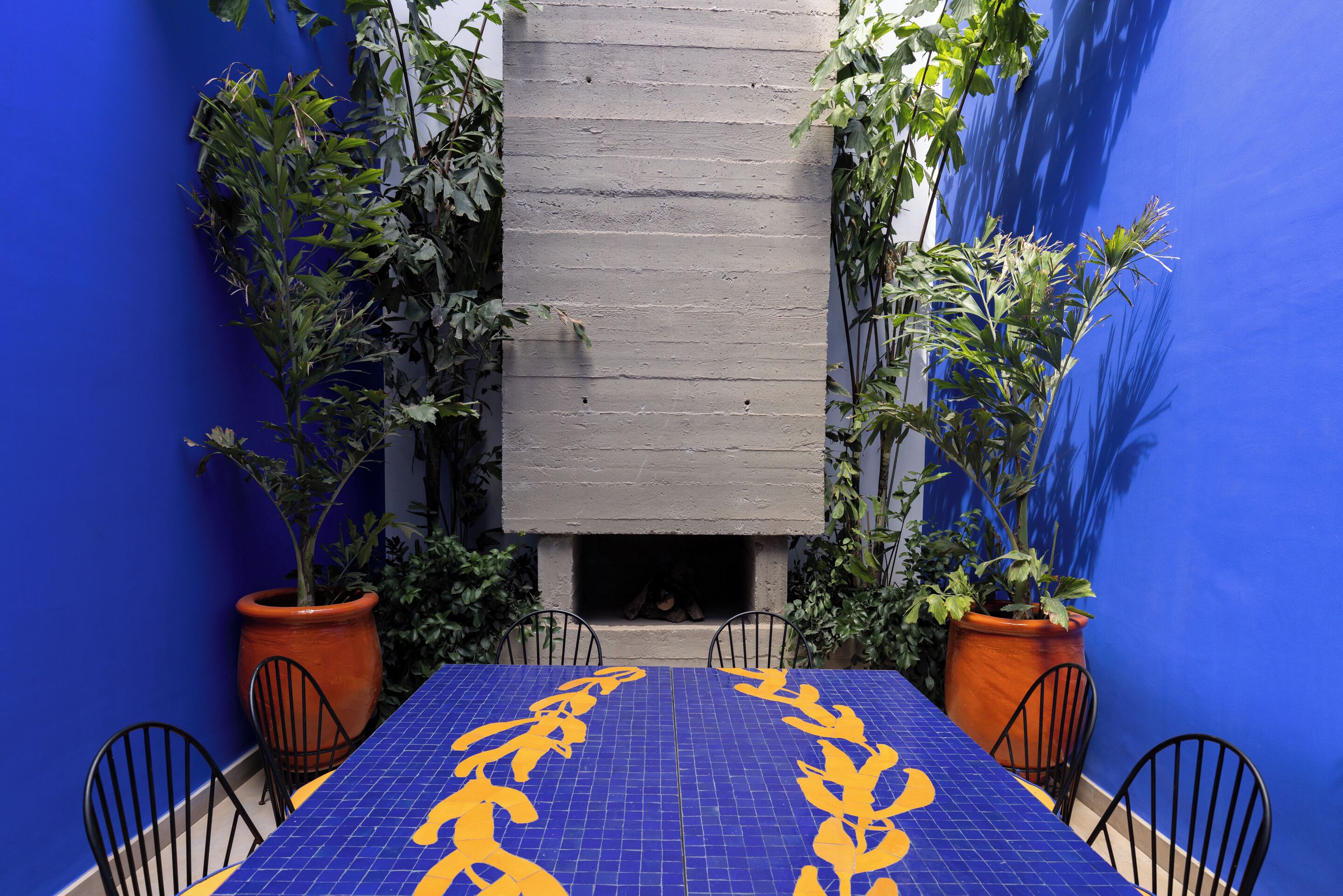 A dining area with a blue tiled table and chairs, surrounded by potted plants and a concrete wall.