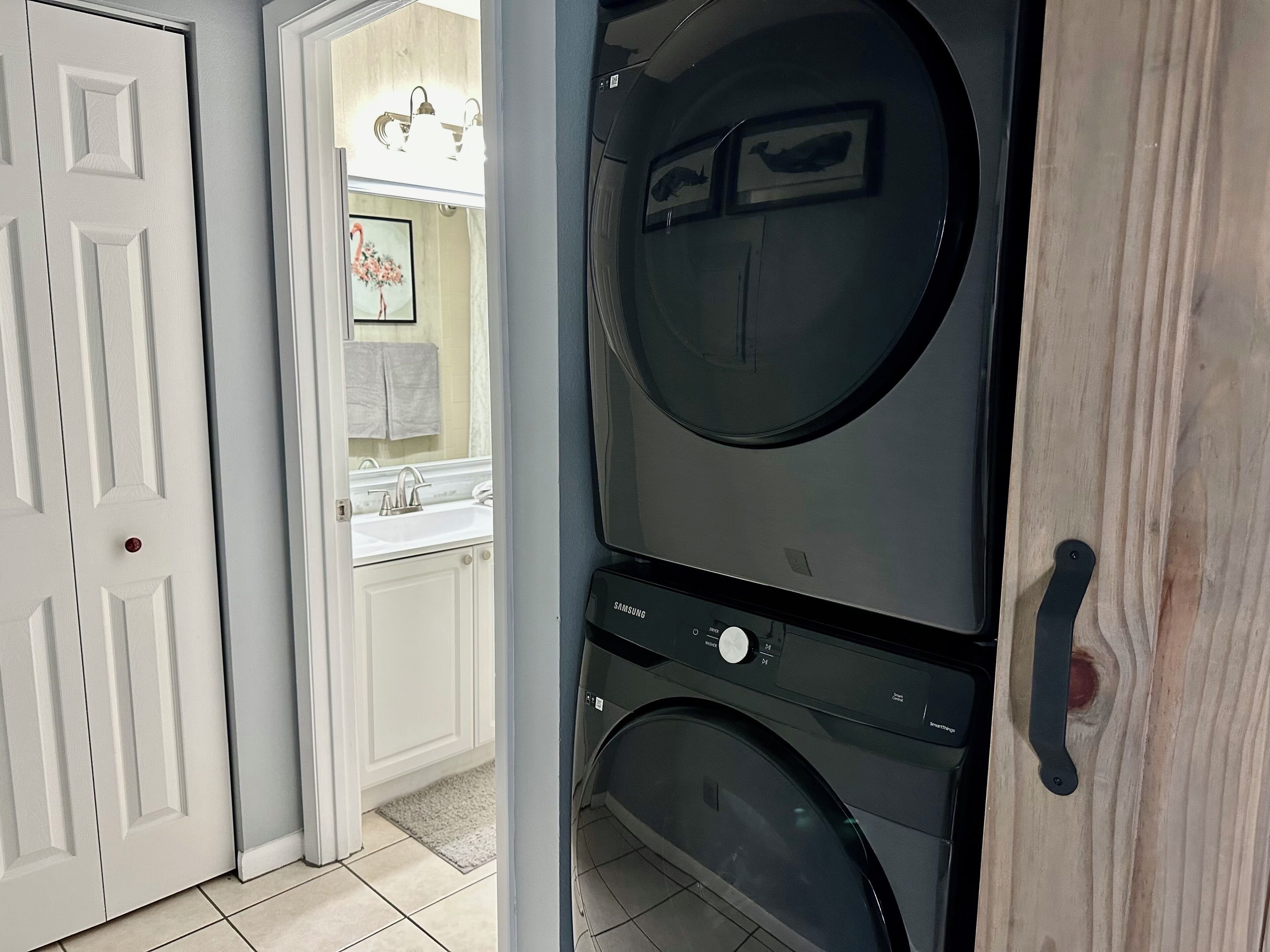 Uma lavanderia com máquina de lavar e secar frontal, uma porta branca e uma parte visível de um banheiro com pia e porta-toalhas.