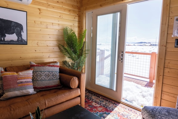 Un interno in legno con un divano in pelle marrone, cuscini decorati, una pianta in vaso e un'ampia finestra che si affaccia su un paesaggio innevato.