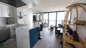 Een moderne keuken met witte kasten, een blauwe accentmuur en een groot eiland. Er is een zithoek met een tafel en stoelen, en een houten plank met decoratieve objecten.