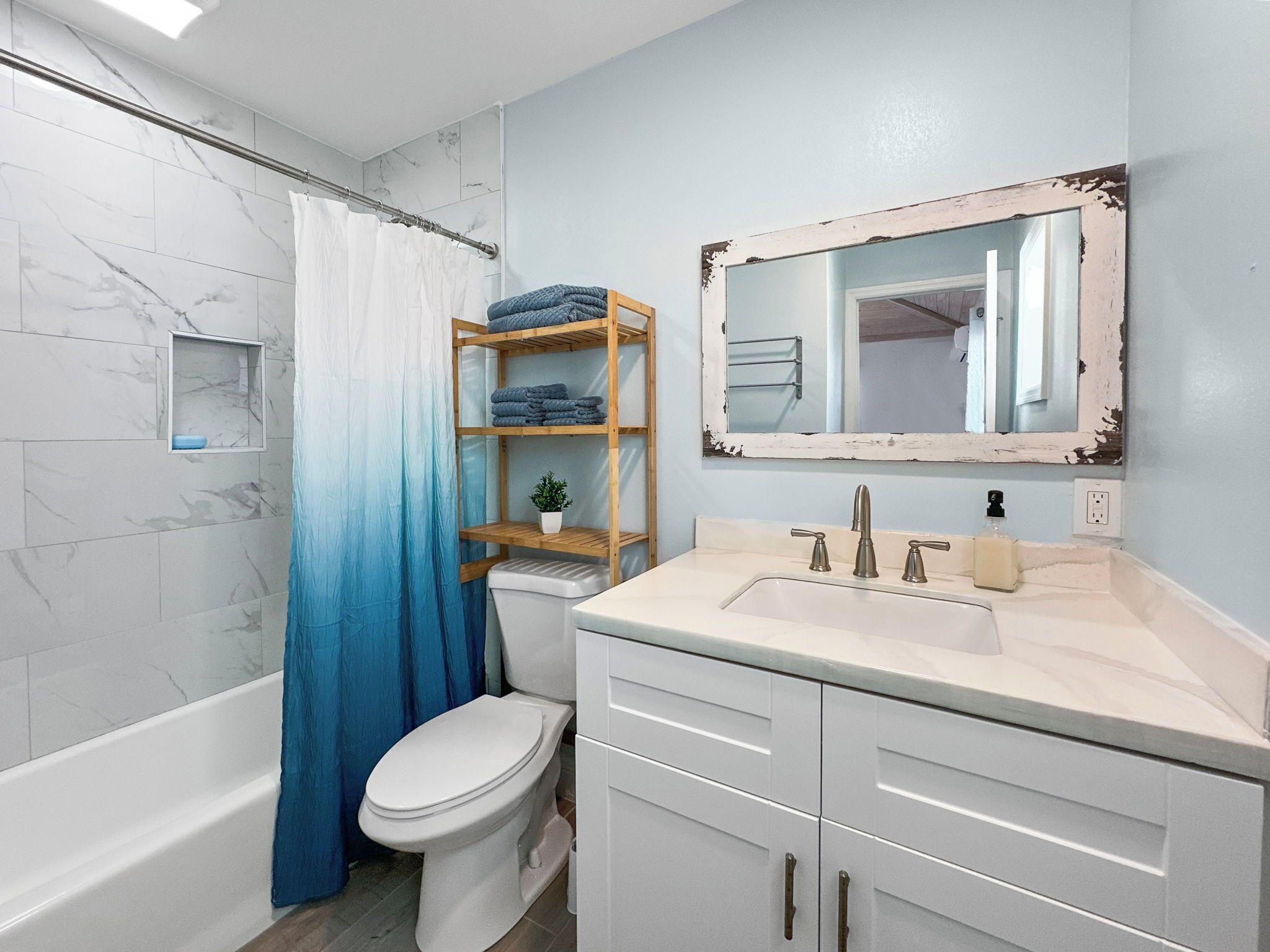 A bathroom with a white toilet, a bathtub, a shower curtain, a wooden shelf with towels, a sink with a mirror, and a small plant.