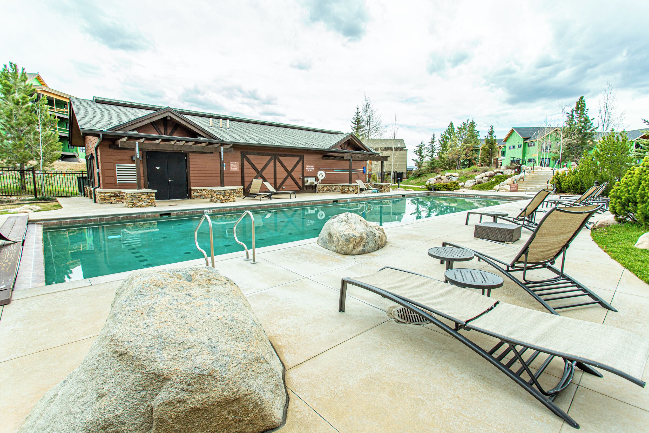 Zona de piscina con sillones reclinables, un edificio de madera y una roca de piedra.
