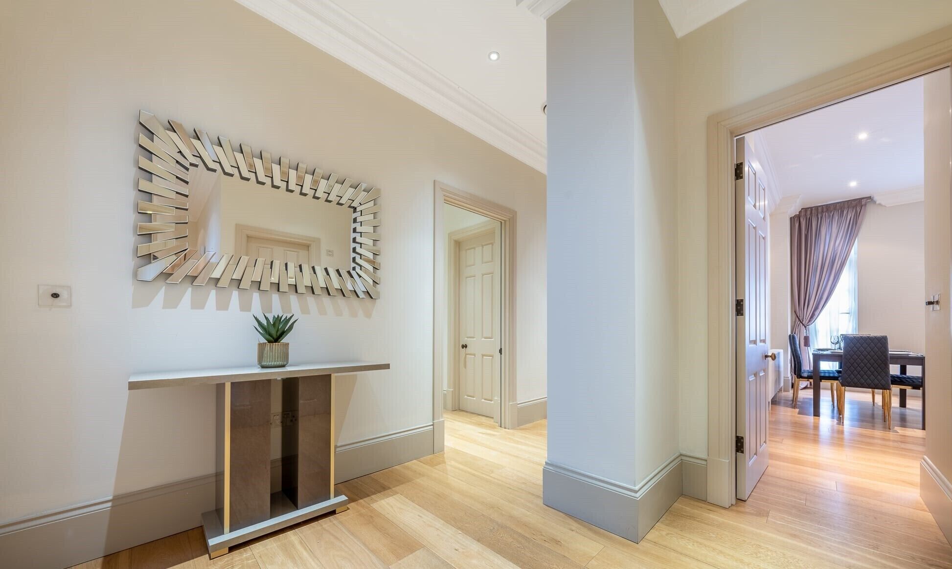 A modern hallway with a decorative mirror, a console table with a plant, and a dining area with a table and chairs.