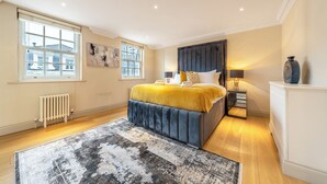 A bedroom with a large bed, a radiator, a window, and a nightstand with a lamp. - The Mayfair Collection (London)