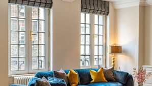 A room with a blue sofa set, a table with a vase of flowers, and a lamp. - The Mayfair Collection (London)