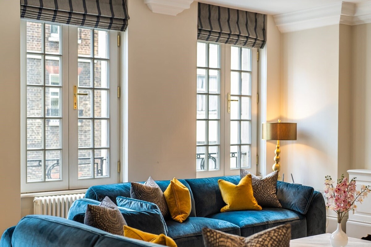 A room with a blue sofa set, a table with a vase of flowers, and a lamp.