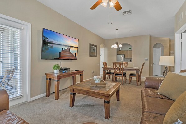 A living room with a wooden coffee table, a brown leather sofa, a dining area with a wooden table and chairs, a kitchen in the background, and a ceiling fan. - Disney 2 miles w/Heated Pool, water park, basketball tennis courts, game room (Kissimmee)