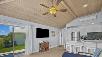 Un vaste espace de vie doté d’un ventilateur de plafond, d’un téléviseur fixé au mur, d’une cuisine avec des meubles blancs et d’une vue sur l’extérieur.