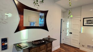 A modern interior with a wooden floor, a dark wooden cabinet, a wall-mounted mirror, and hanging green plants. - Cozy, custom bungalow in the heart of the Sante Fe Art District! (Denver)
