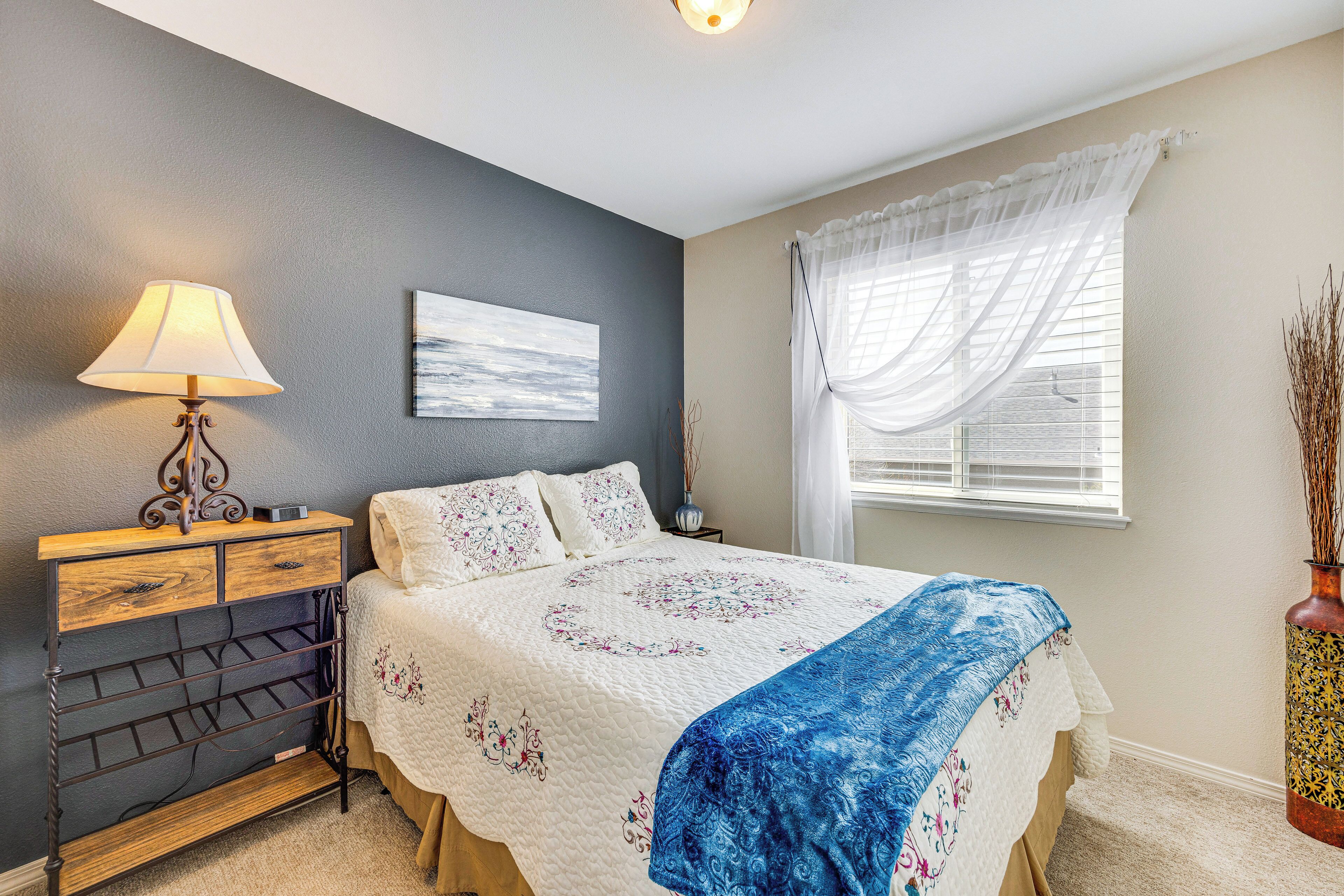 A bedroom with a bed, bedside table, lamp, artwork, window with sheer curtains, and a vase with dried plants.
