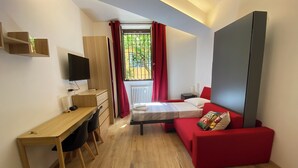 A compact room with a wooden desk, a bed with red bedding, a red sofa, and a large grey cabinet. - Under Trastevere stars - Live like a local on Gianicolo hill! (Roma)