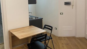 A small, open-plan living space with a wooden dining table and two black chairs, a kitchen area with a modern light fixture, and a white door. - Under Trastevere stars - Live like a local on Gianicolo hill! (Roma)