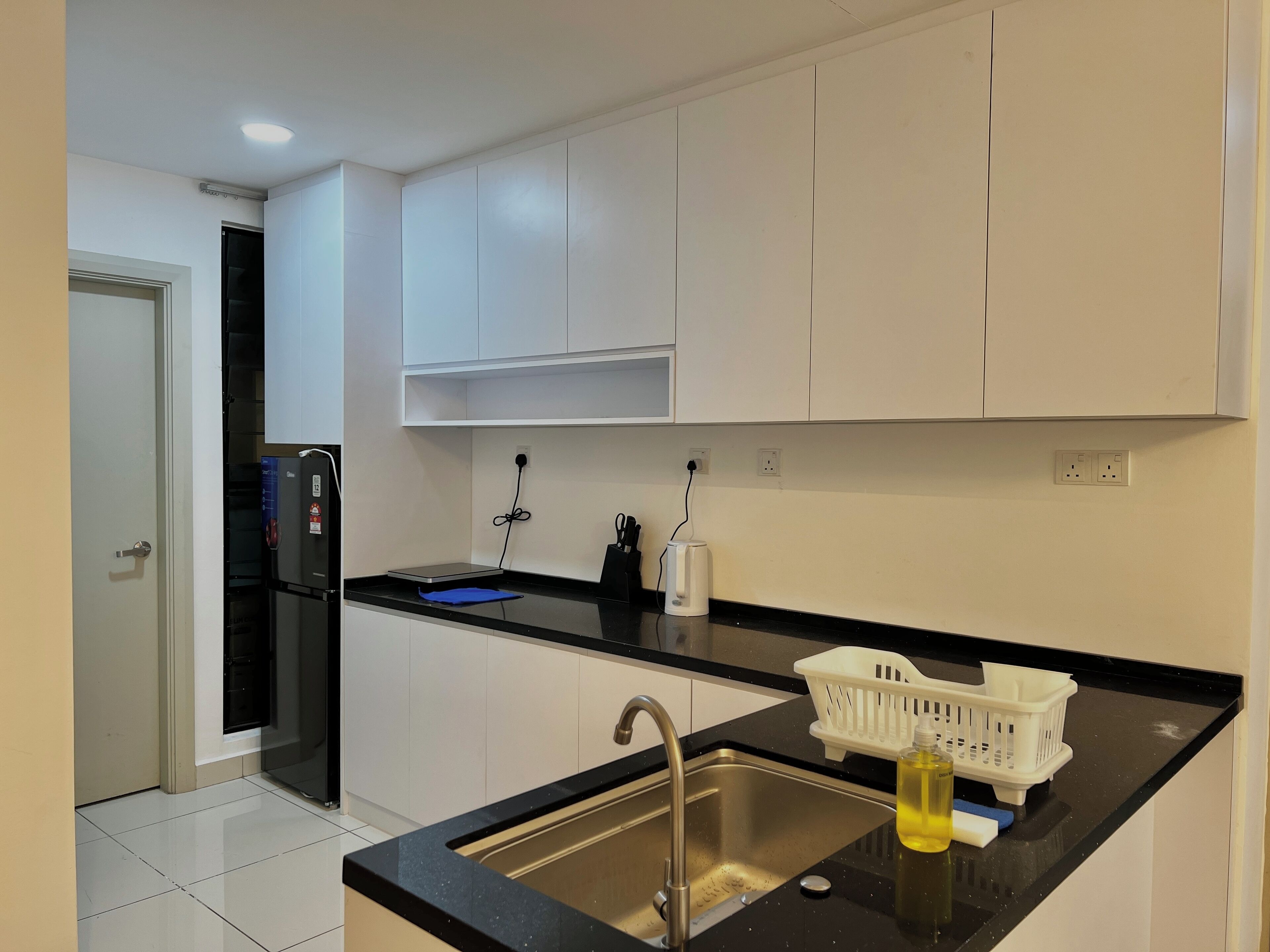 A modern kitchen with white cabinets, a black countertop, a stainless steel sink, and a small white basket with a bottle.