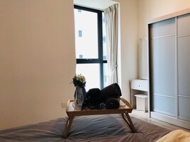 A bedroom with a bed, a bedside table, a window with curtains, and a view of buildings outside.
