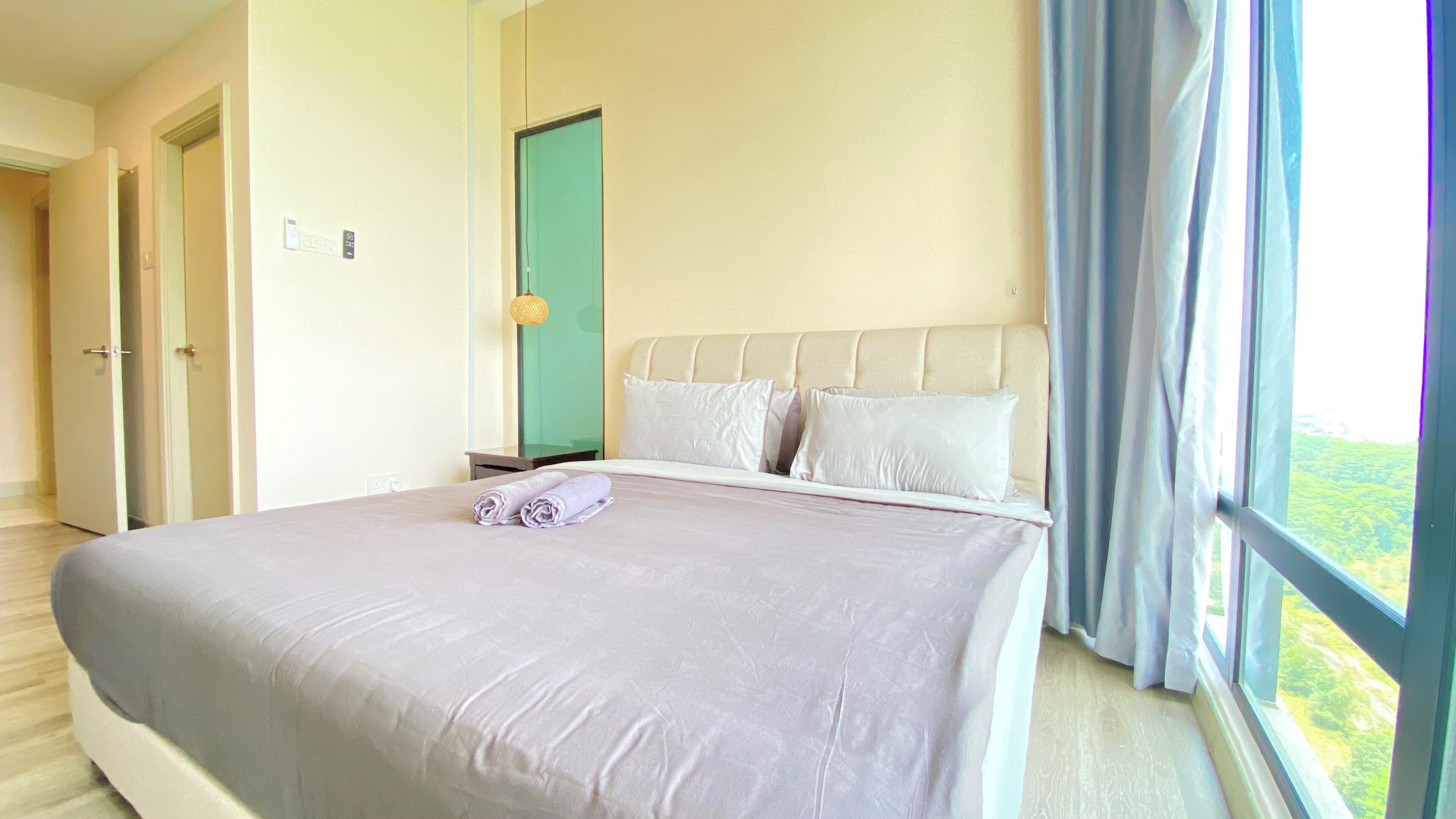 A hotel room with a large bed, a nightstand, a green door, and a view of greenery through the window.