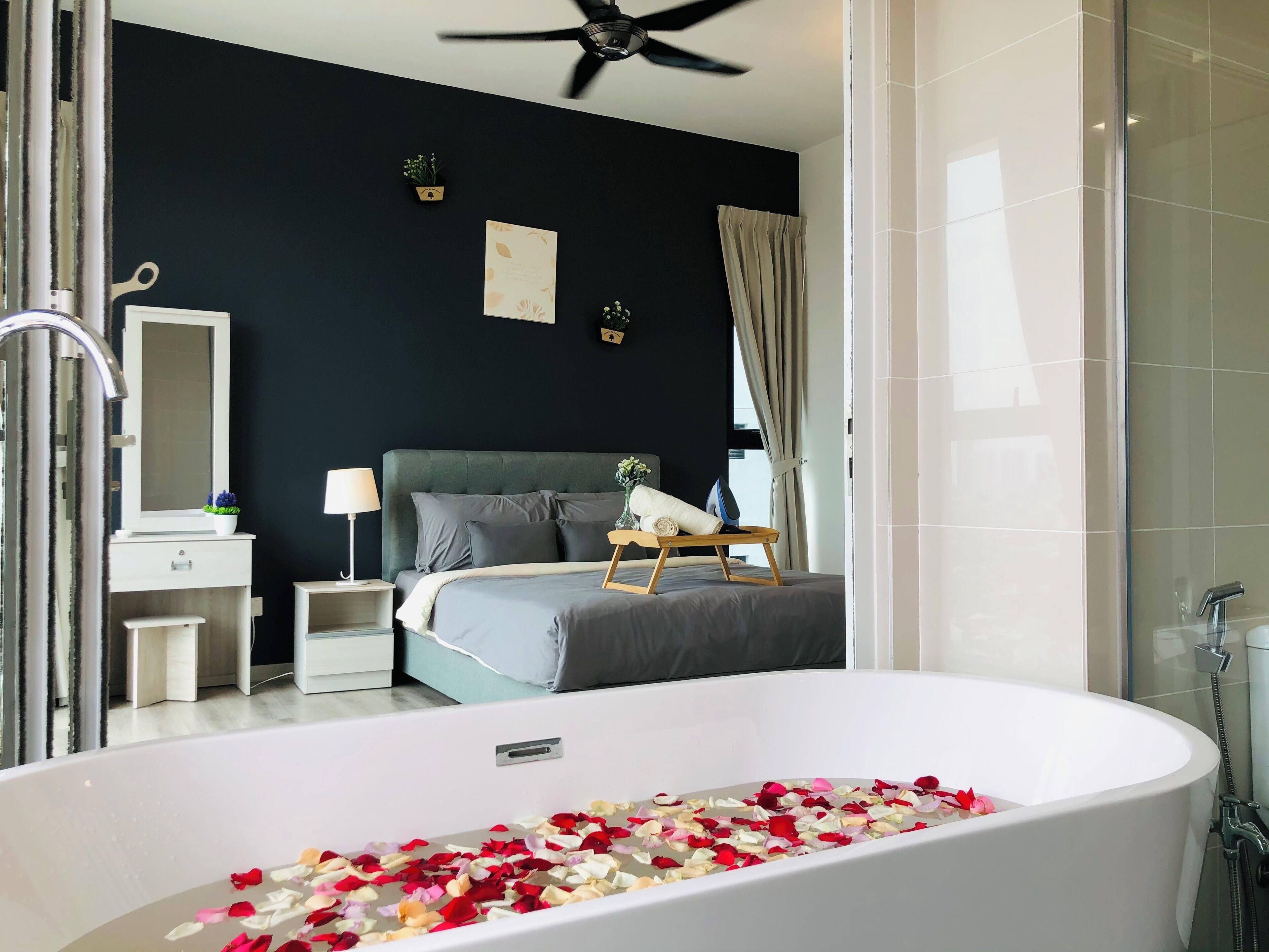 Un dormitorio moderno con una bañera grande llena de pétalos de rosa, una cama con ropa de cama gris, una mesita de noche con una lámpara y una pequeña planta en maceta en la mesita de noche.