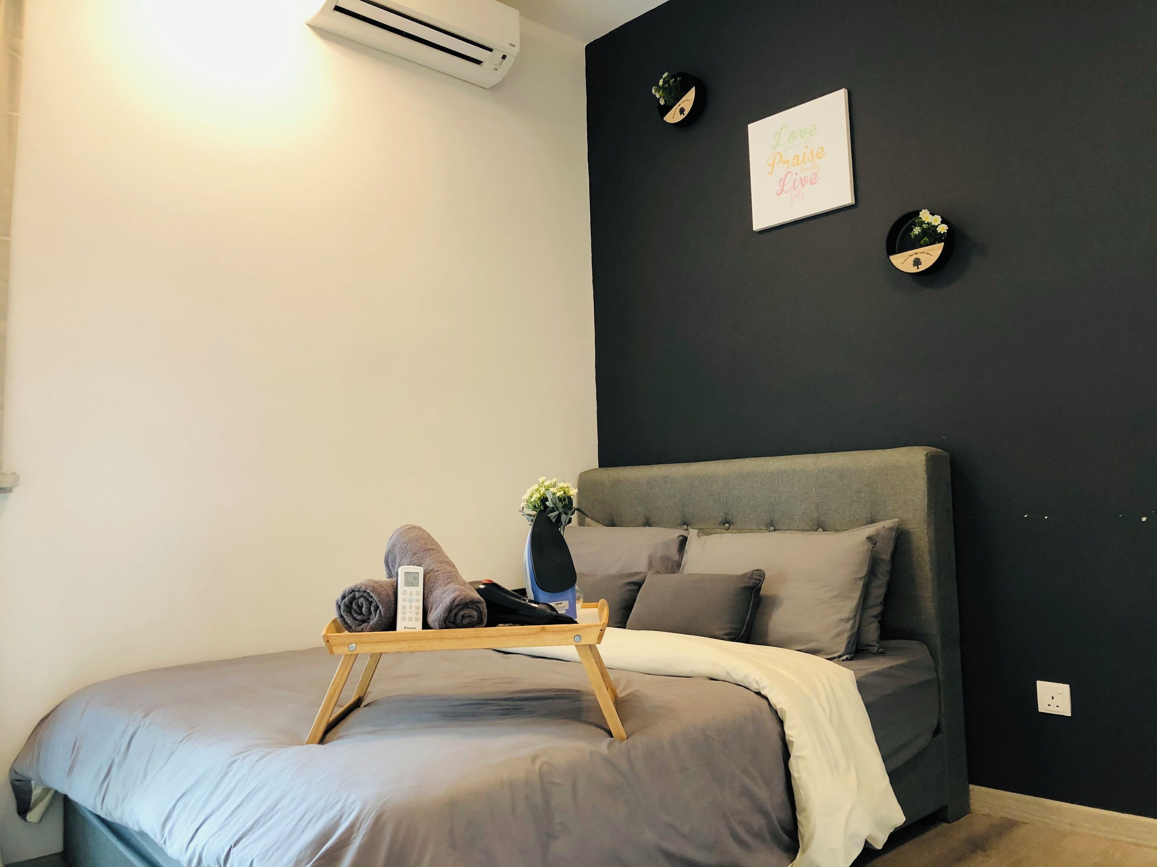 A modern bedroom with a grey bed, a bedside table with a towel and a phone, a wall-mounted air conditioner, and two wall decorations.