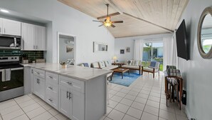 Cocina moderna con armarios blancos, horno empotrado y microondas. Cuenta con un área de comedor con sofá y mesa de centro. La habitación tiene un ventilador de techo y un ventanal con vistas al exterior.