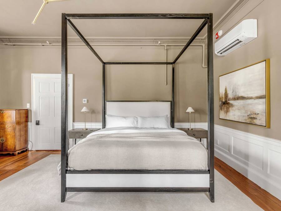 Una cama con dosel, ropa de cama en tonos neutros, una mesita de noche con una lámpara, un aparador de madera y un cuadro en la pared.