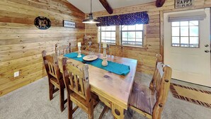 Een houten hut interieur met een eettafel, stoelen, een kroonluchter en een deur met een bordje.