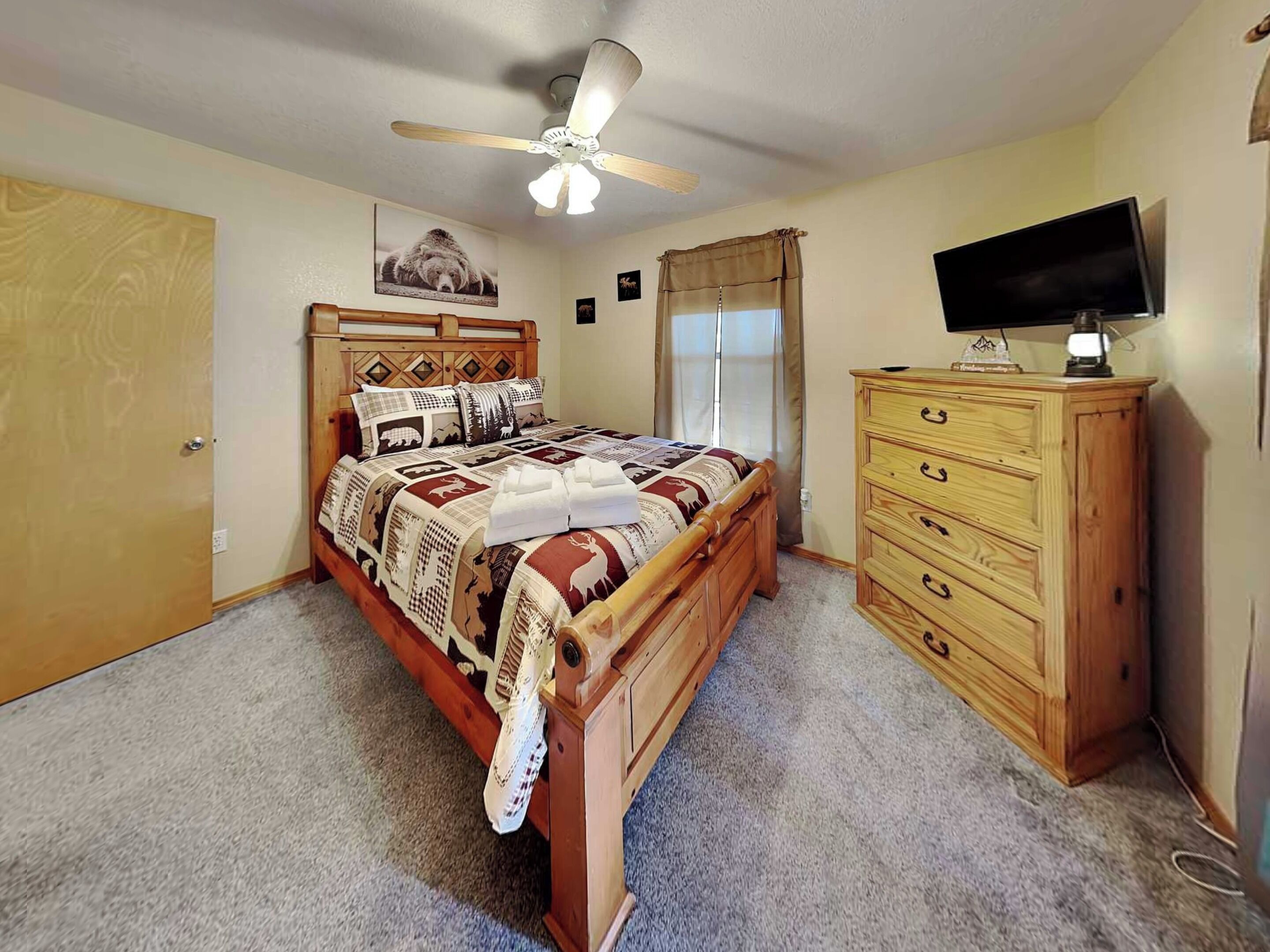 A bedroom with a wooden bed, a ceiling fan, a TV on a wooden dresser, and a window with curtains.