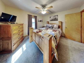 A bedroom with a bed, a wooden dresser, a television on a stand, a ceiling fan, and a window with curtains.