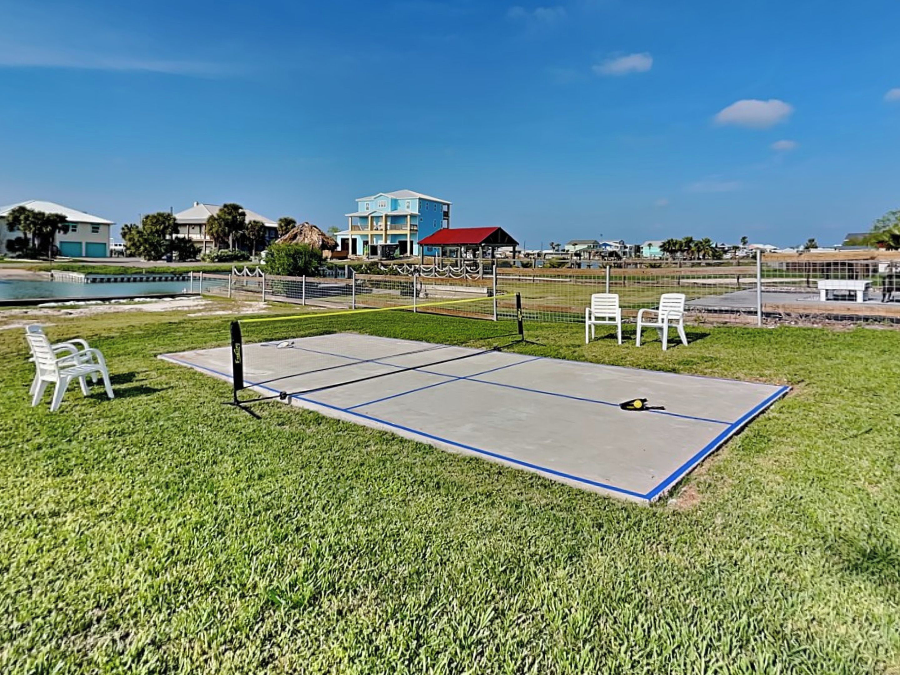 Una cancha de tenis con una red, rodeada de césped, sillas blancas y un edificio al fondo.