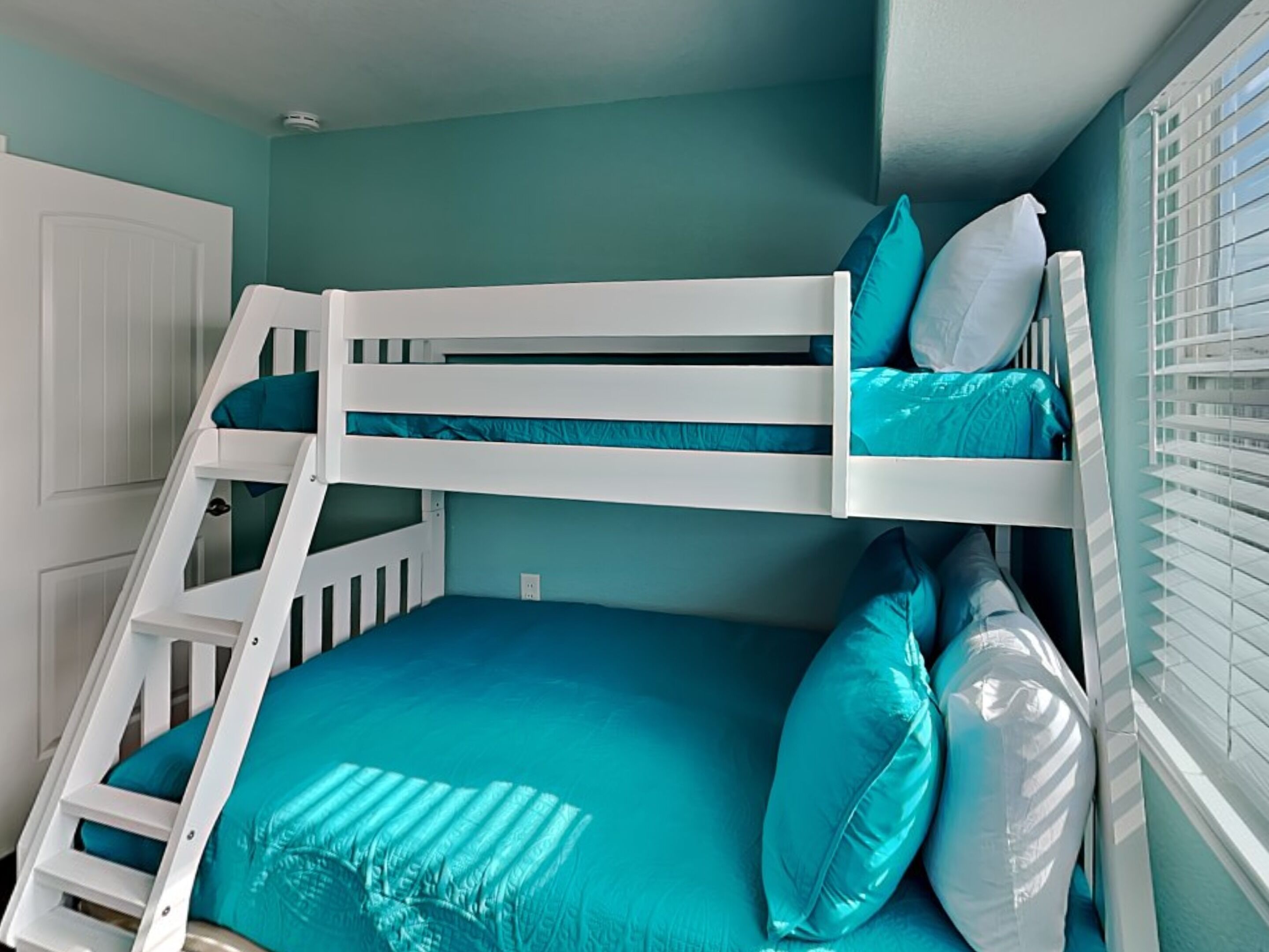 Een stapelbed met een ladder, blauwgroene beddengoed en witte kussens.