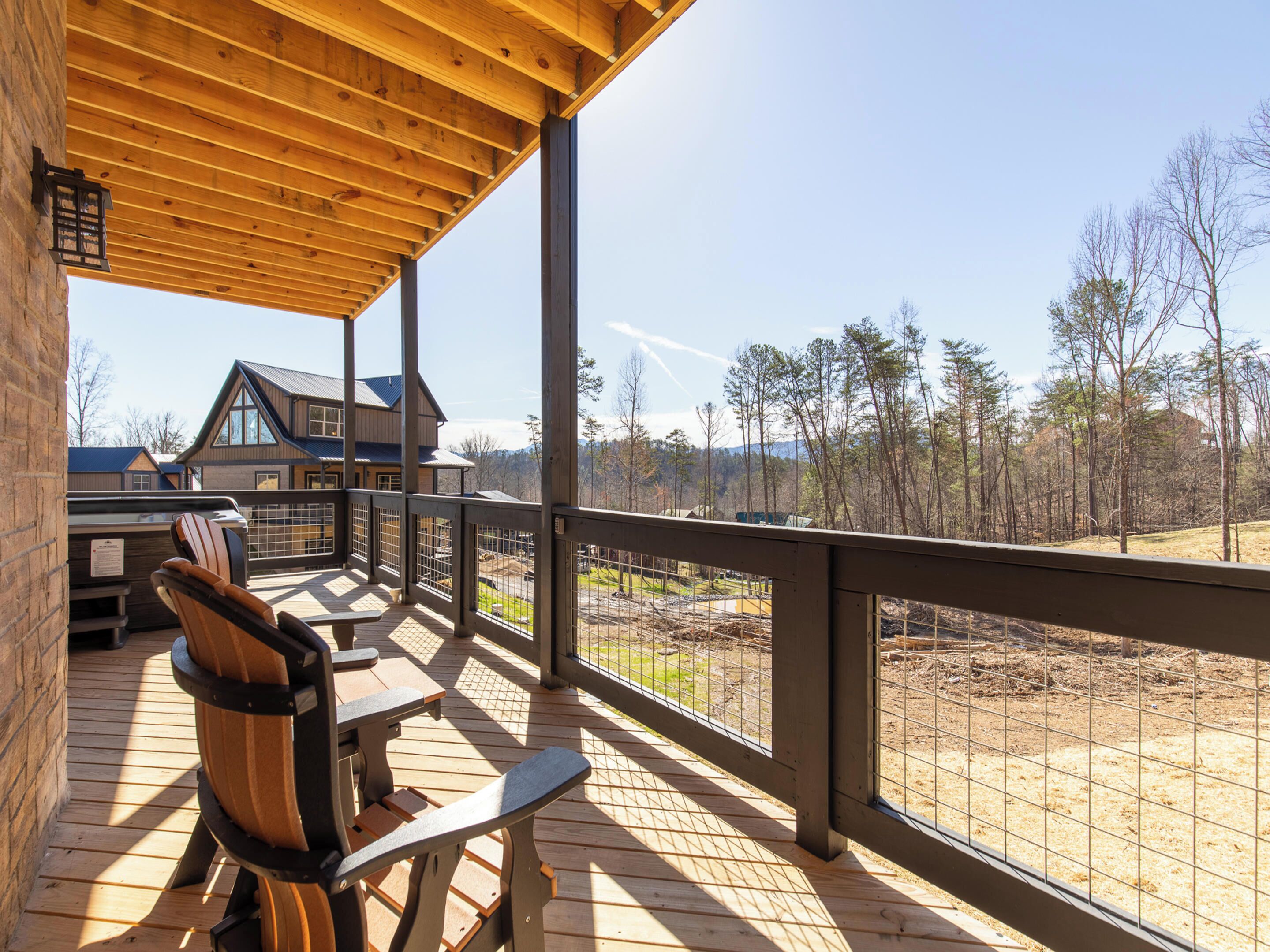 A wooden deck with chairs and a table, overlooking a fenced area with trees and a building in the background.