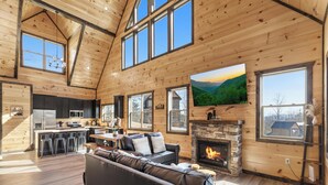 A spacious living area with a fireplace, a large TV mounted on the wall, and a kitchen in the background.