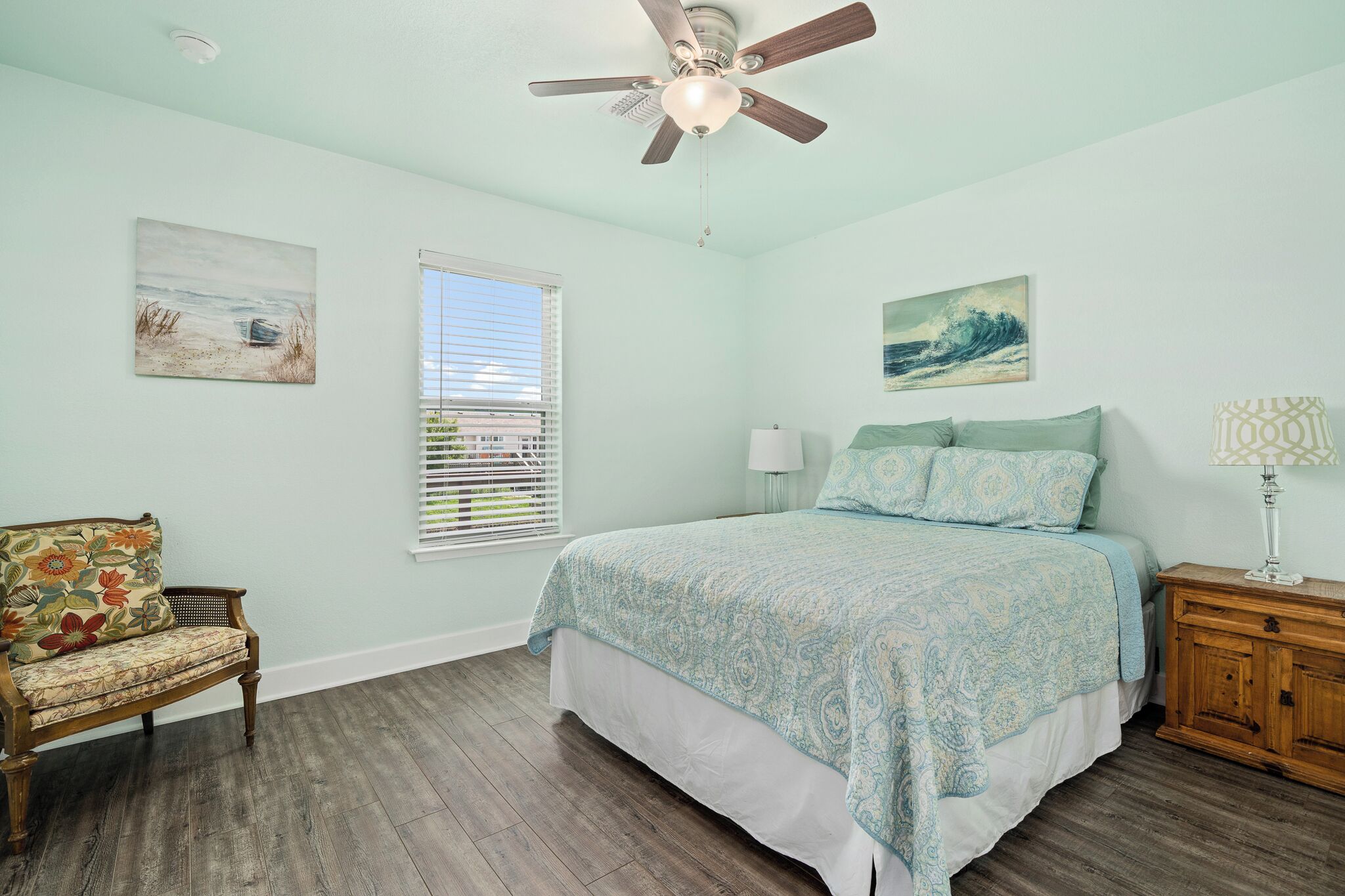 A bedroom with a bed, a chair, a nightstand, a ceiling fan, and two paintings on the walls.