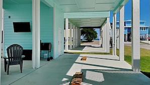 A covered porch with a view of a blue building and a tree in the distance. - Come And Catch It by Miss Kitty’s (Rockport)