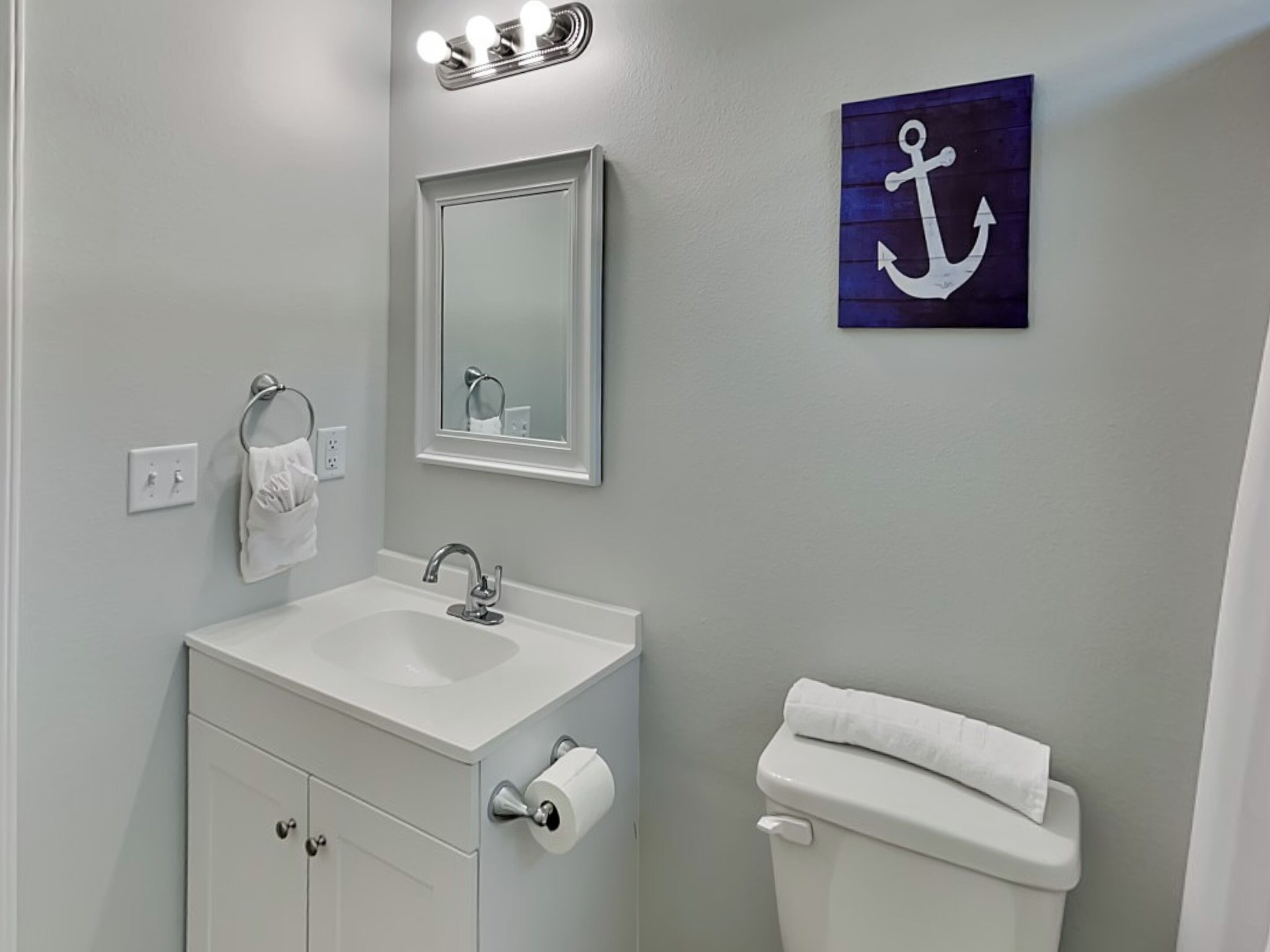 A bathroom with a white sink, mirror, and toilet. There is a towel rack with a towel, a roll of toilet paper, and a wall-mounted light fixture above the mirror.