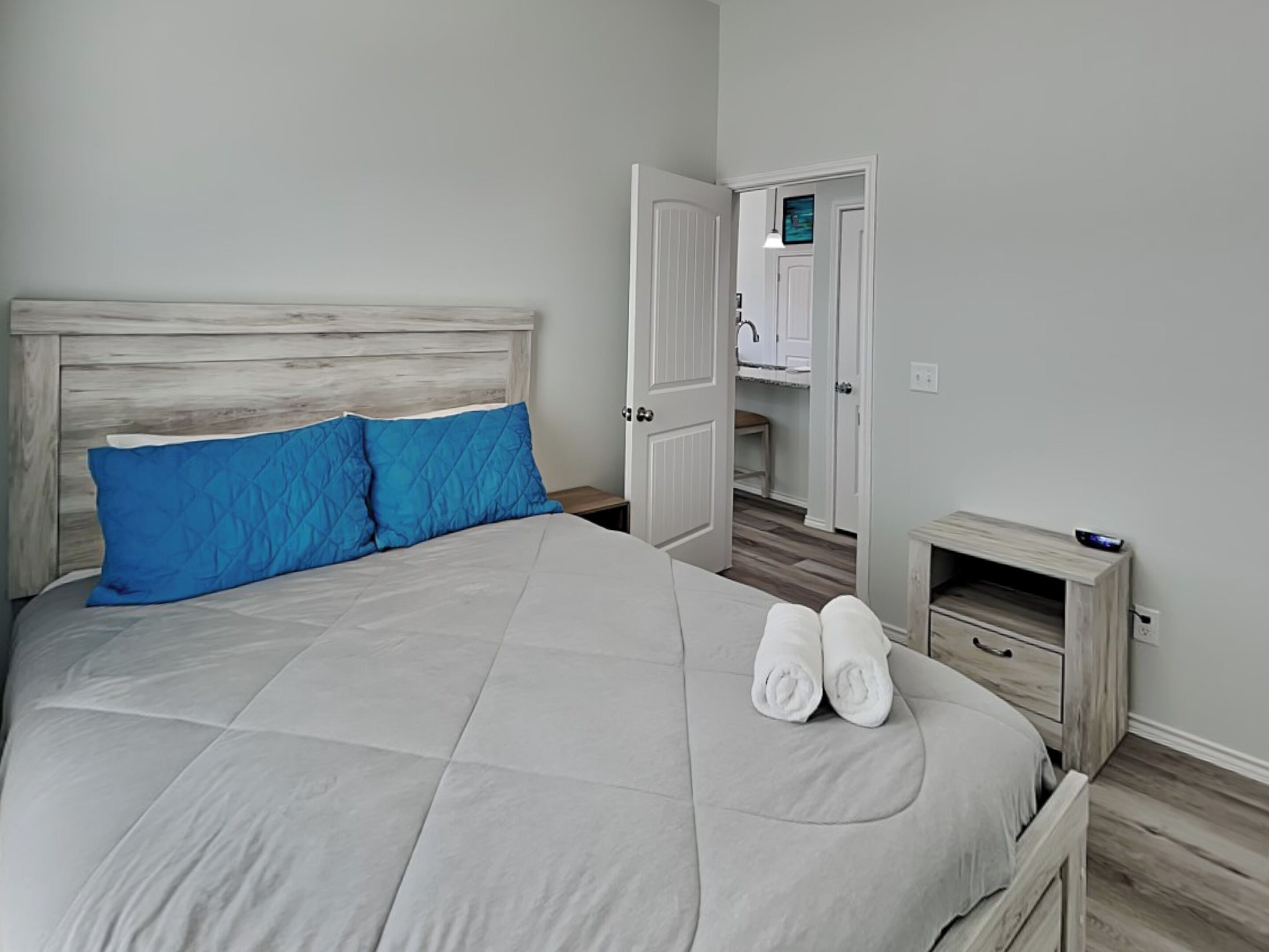 Un lit bien fait, avec une tête de lit matelassée, une paire de serviettes pliées et une petite table de chevet sur laquelle se trouve une télécommande.