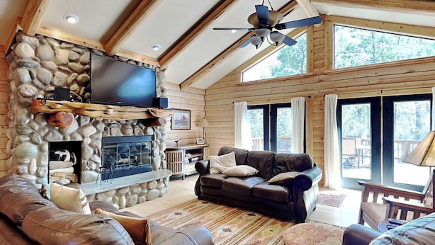 A log cabin interior with a stone fireplace, leather sofas, a flat-screen TV, and large windows. - Double Eagle Manor by Ruidoso Vacation Rentals (Alto)