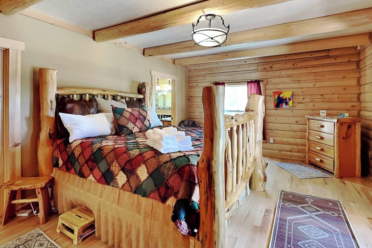 A cozy wooden cabin bedroom with a large bed, a wooden dresser, and a small stool.