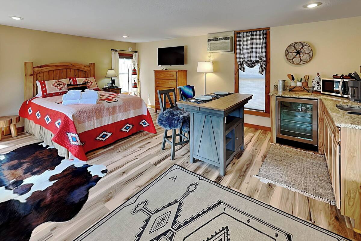 A bedroom with a large bed, a desk, a television, and a kitchenette.
