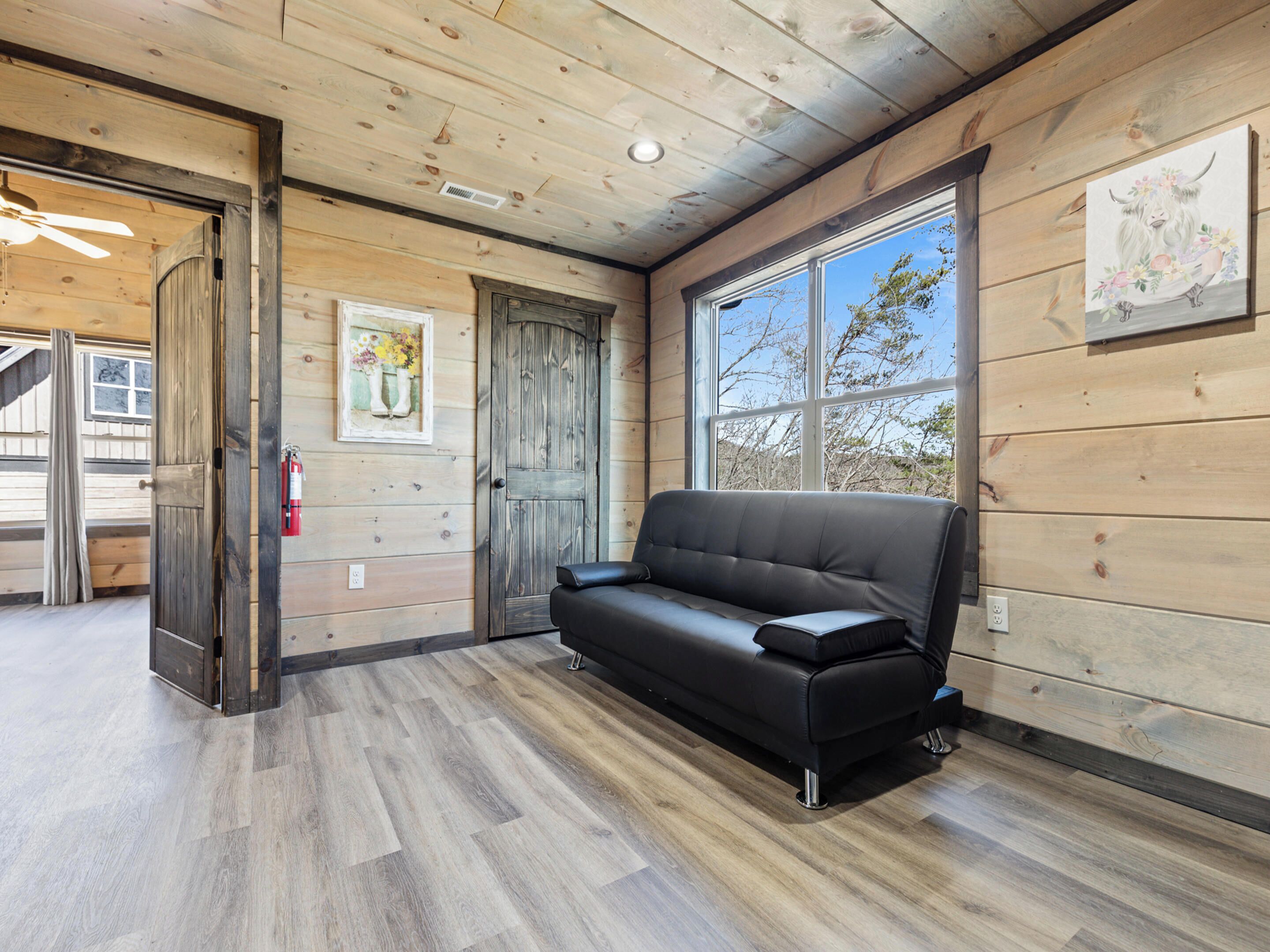 A wooden interior with a black leather sofa, a wooden door, and a window with a view of trees.