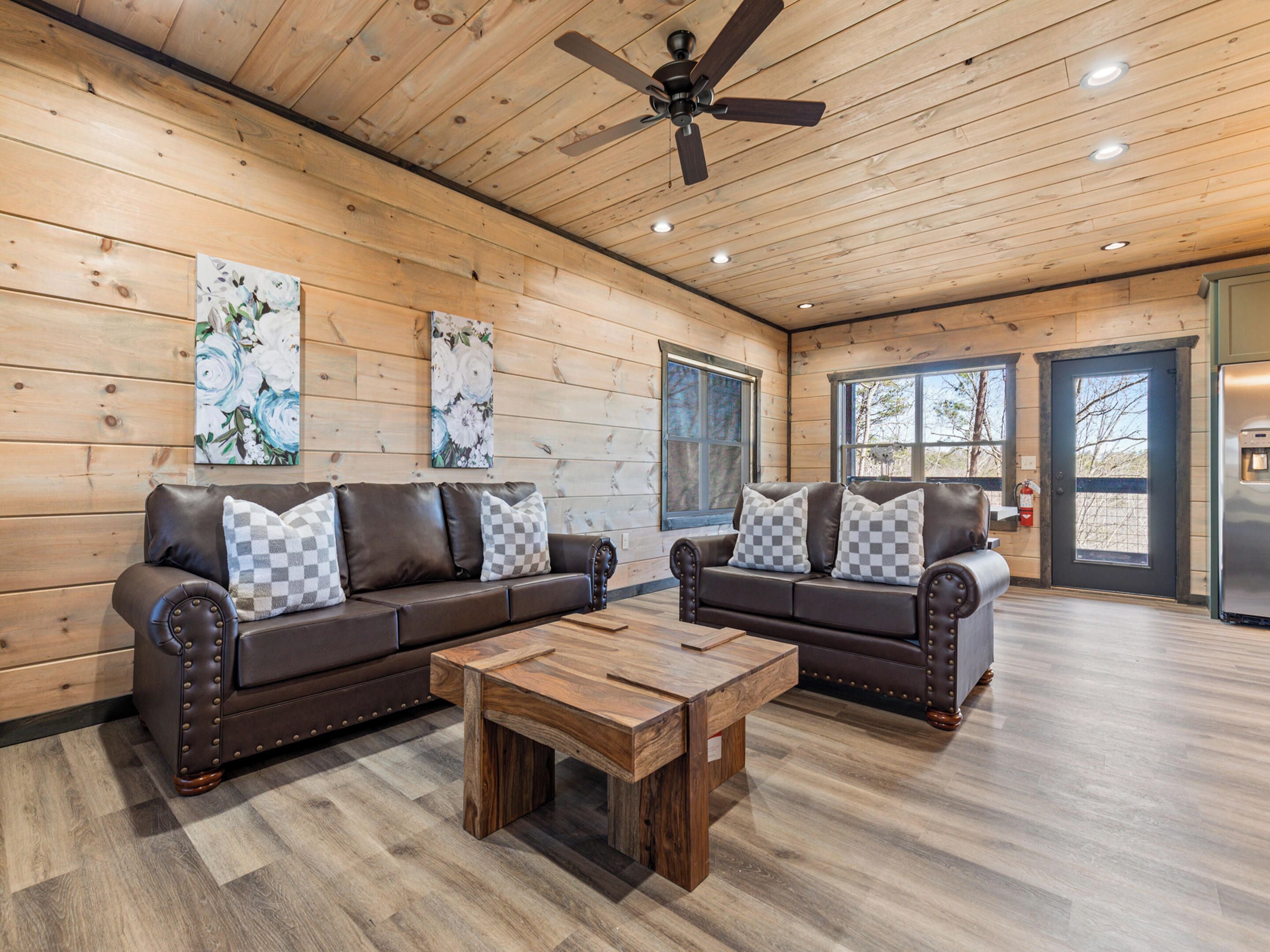 A spacious living room with two leather sofas, a wooden coffee table, wooden walls, a ceiling fan, and large windows.