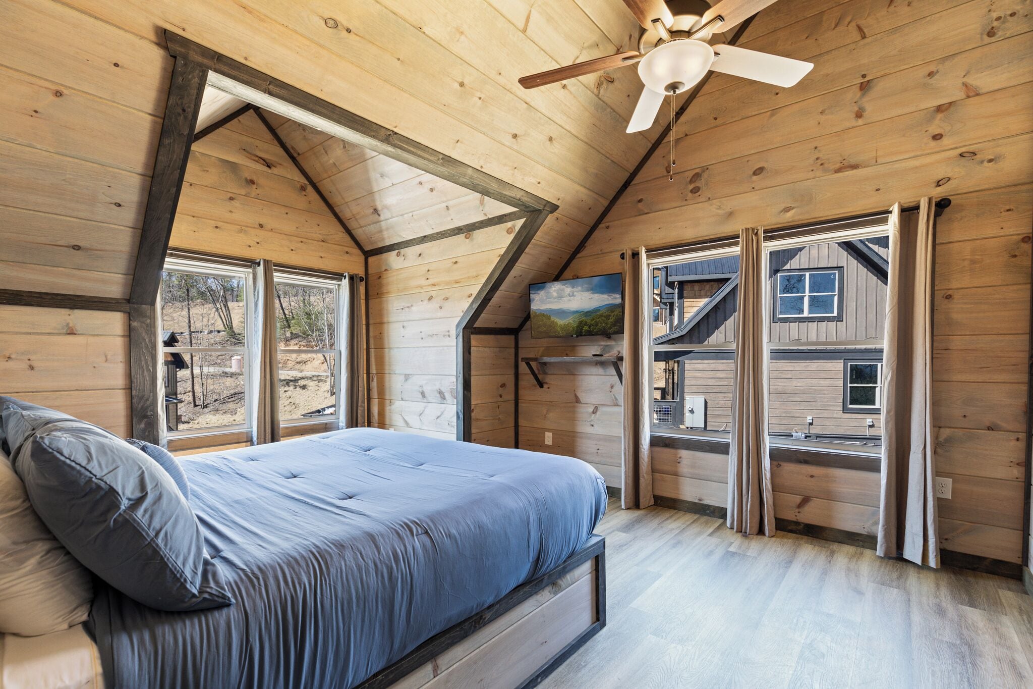 A bedroom with a bed, wooden walls, a ceiling fan, and large windows.