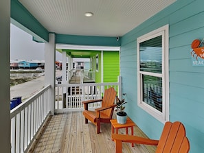 A porch with wooden flooring, two wooden Adirondack chairs, a small table with a potted plant, and a sign indicating a beach location.