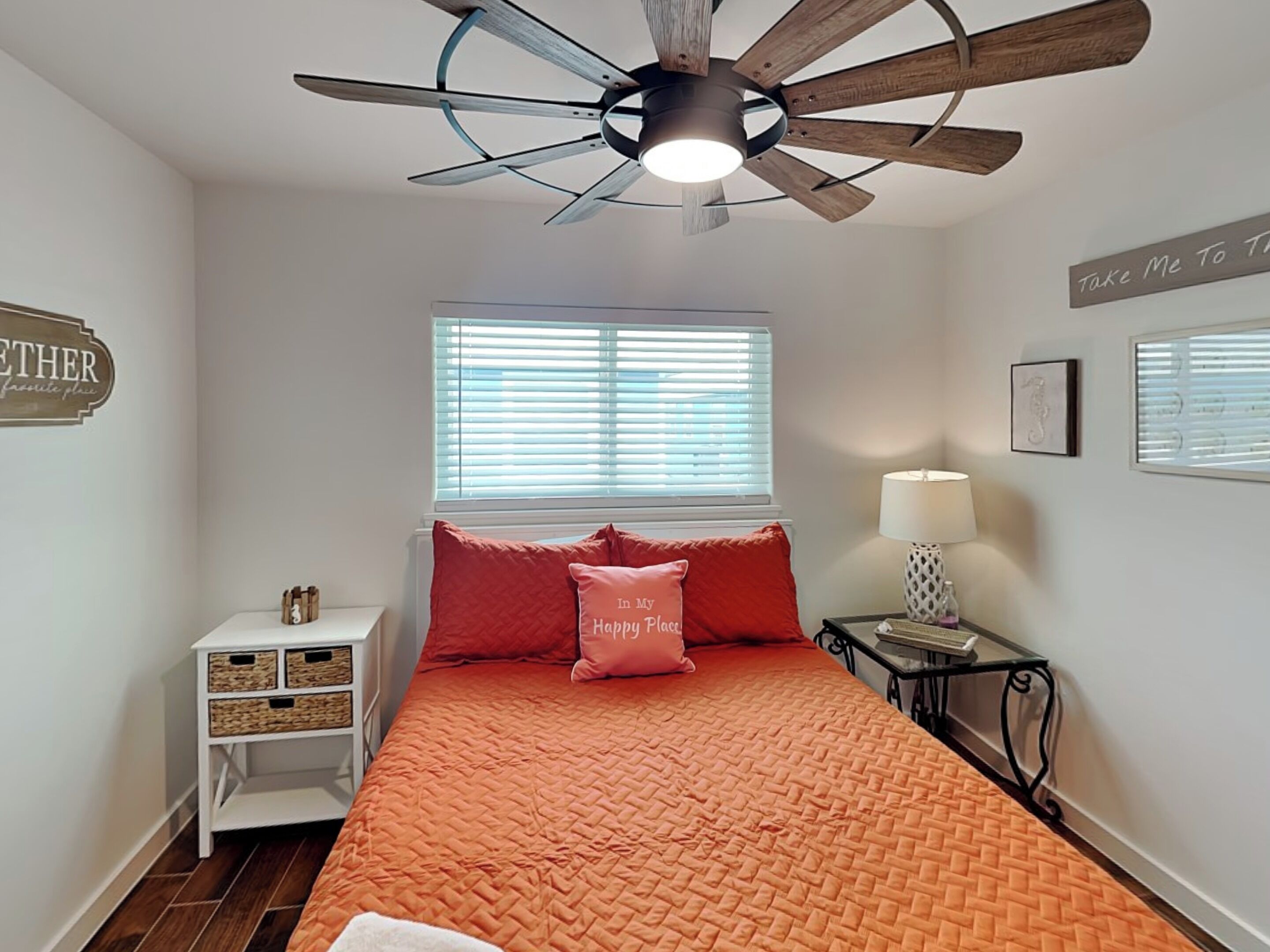 A bedroom with a bed, a ceiling fan, a window with blinds, a bedside table with a lamp, and a wall decoration.