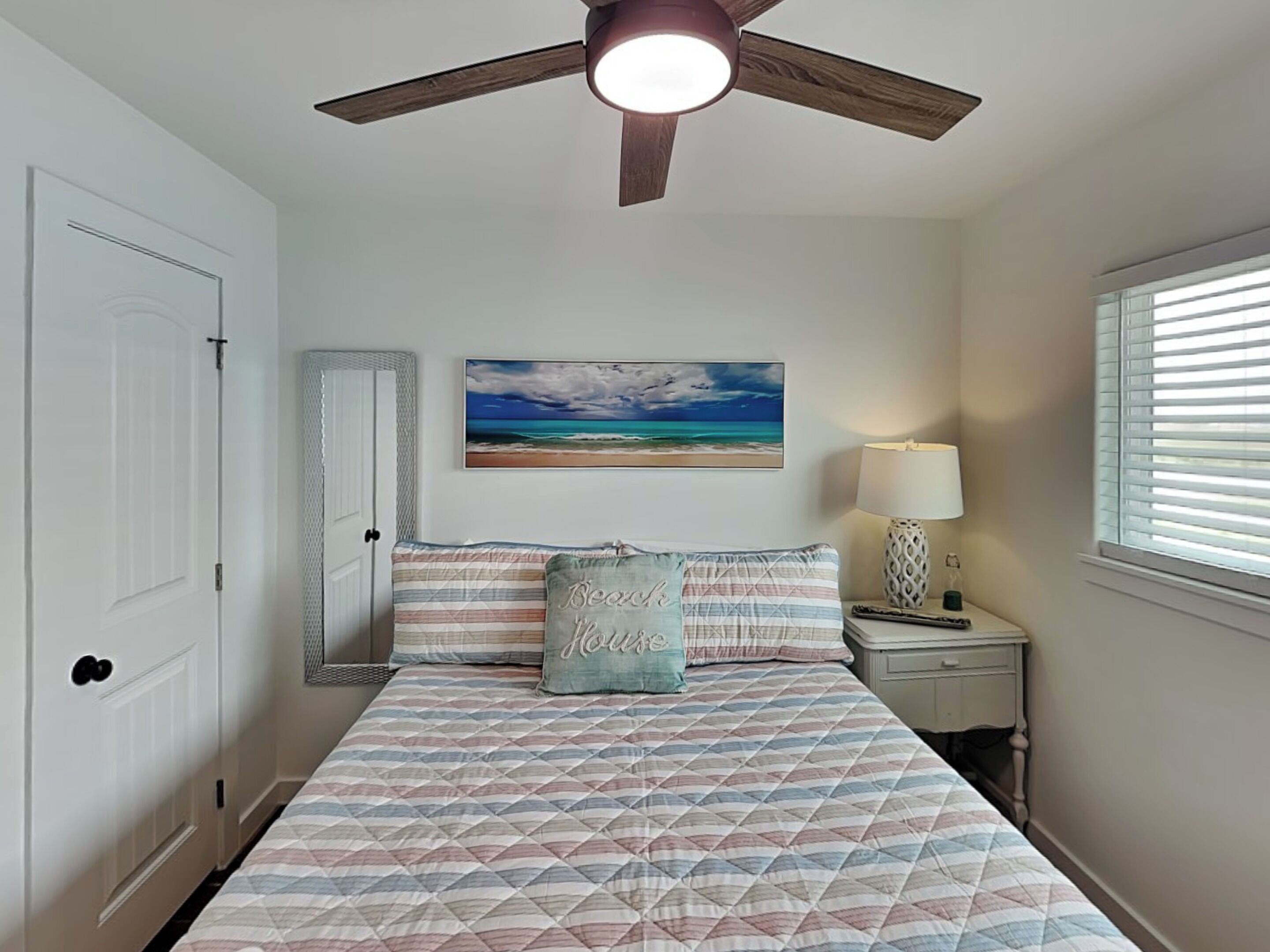 Une chambre à coucher avec un lit, un ventilateur de plafond, une table de chevet avec une lampe, une fenêtre avec des stores et un tableau sur le thème de la plage accroché au mur.