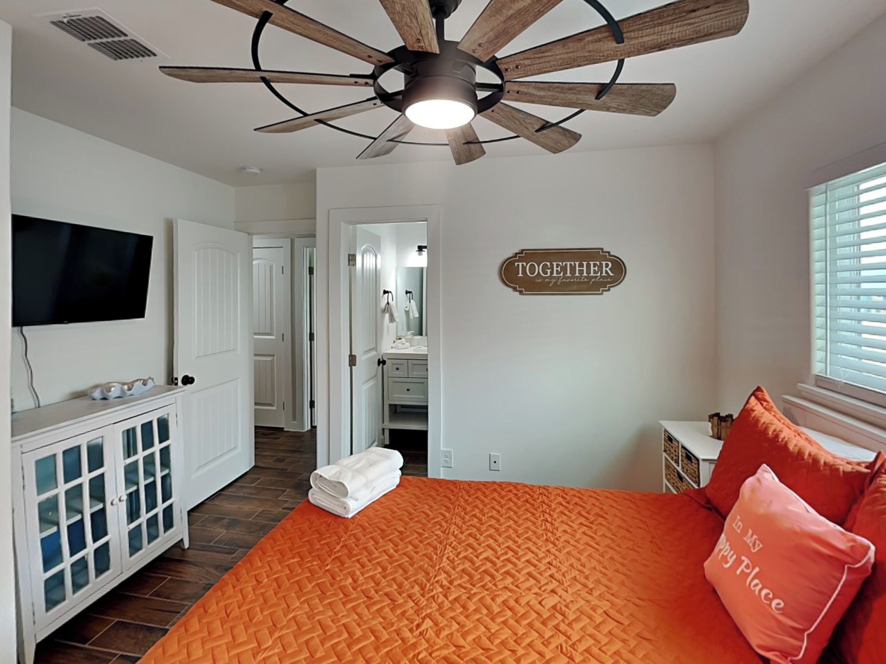 Een slaapkamer met een plafondventilator, een bed met oranje beddengoed, een televisie, een dressoir en een bord met de tekst 'TOGETHER'.
