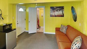 Quarto com ventilador de teto, micro-ondas, sofá com almofadas estampadas e porta que dá para outro cômodo.