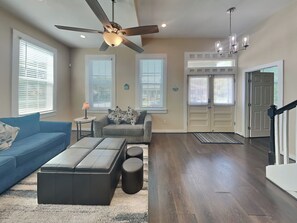A spacious living room with a blue sofa, a grey armchair, and a black ottoman. There is a ceiling fan, a chandelier, and a door with a glass panel. - Seastar BRM by Ryson Vacation Rentals (Galveston)