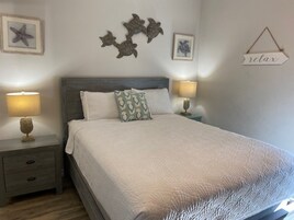 Una cama bien tendida con colcha blanca, dos mesitas de noche con lámparas y decoración en la pared con motivos de estrellas de mar y tortugas.