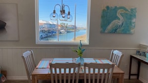 Un comedor con una mesa y sillas de madera, vistas a un puerto deportivo a través de un ventanal grande, y un cuadro de un pájaro en la pared.