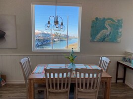 Un comedor con una mesa y sillas de madera, vistas a un puerto deportivo a través de un ventanal grande, y un cuadro de un pájaro en la pared.