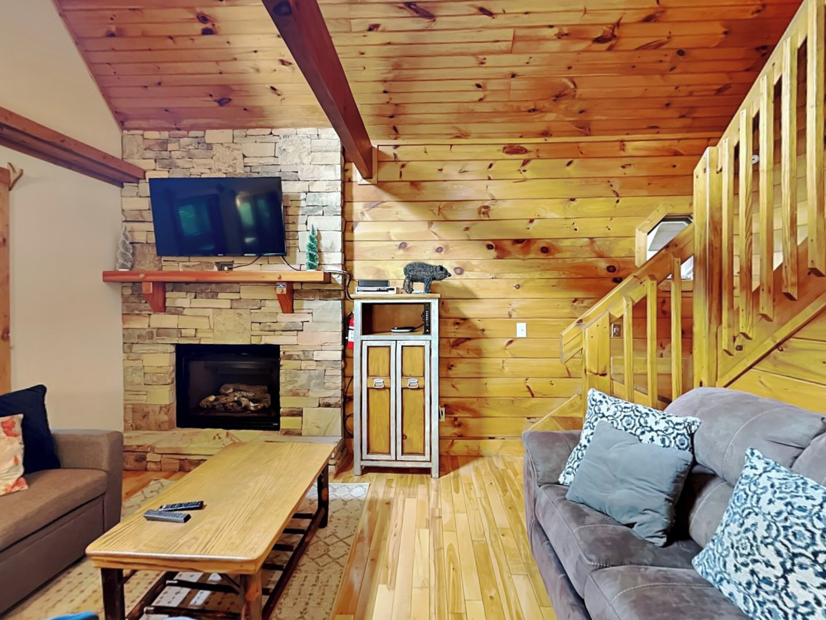 A cozy living room with a stone fireplace, wooden beams, a television, and a sofa with patterned cushions.